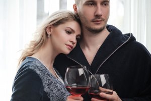 A couple, a man and a woman in bathrobes drink red wine from large glass goblets in the apartment. Kiss and hug.